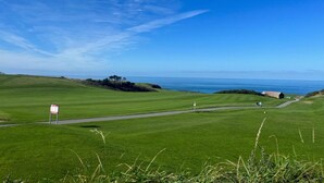Ein Golfplatz mit freiem Blick auf den Ozean, eine Flagge auf einem Fahnenmast und ein Gebäude in der Ferne.