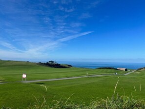 Ein Golfplatz mit freiem Blick auf den Ozean, eine Flagge auf einem Fahnenmast und ein Gebäude in der Ferne.