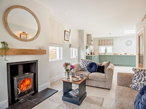 A living room with a fireplace, a sofa, a coffee table, and a kitchen in the background. - 3 Bed in Pwllheli (oc-95040) (Morfa Nefyn)