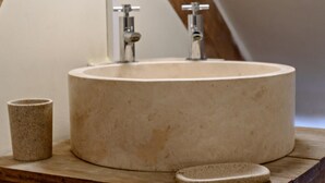 Een ronde beige wastafel met twee kraanwerkjes, een klein beige kopje en een stenen zeepbakje op een houten oppervlak.