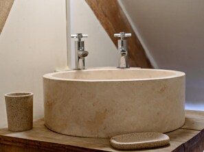 Ein rundes, beiges Waschbecken mit zwei Wasserhähnen, eine kleine, beige Tasse und eine Seifenschale aus Stein auf einer Holzoberfläche.