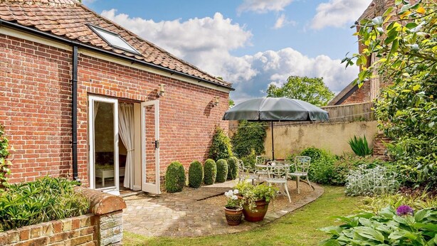 Een bakstenen huis met een terras, een deur, en een tuin met bloempotten en een tafel met stoelen.