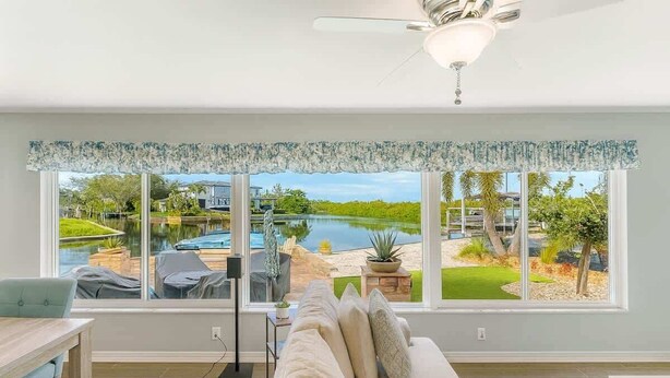 Habitación con ventana grande que da a una piscina y follaje, ventilador de techo y sofá con cojines.