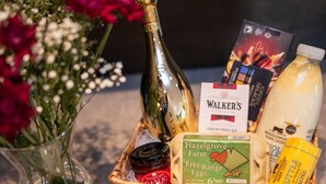 Ein Korbwägelchen mit einer Weinflasche, einer Schachtel Shortbread, einem Glas mit Bio-Eiern vom Bauernhof, einer Butterflasche und einem Buch.