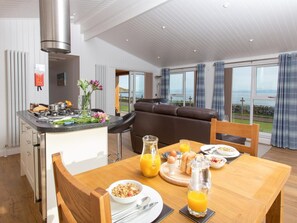A modern kitchen with a central island, dining table with breakfast items, and a view of the outdoors. - 1 Bed in Castle Douglas (oc-64389) (Auchencairn)
