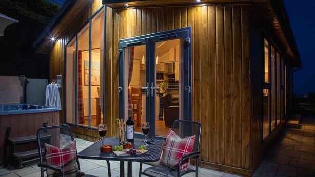 A wooden cabin with a dining area, a hot tub, and a kitchen visible through the glass doors. - 1 Bed in Castle Douglas (oc-rasc2) (Auchencairn)