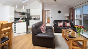 A modern kitchen with white cabinets, a dining area with a wooden table and chairs, a living room with a leather sofa, and a large window offering a view of the outdoors. - 1 Bed in Castle Douglas (oc-rasc4) (Auchencairn)