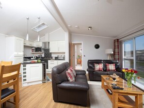 Eine moderne Küche mit weißen Schränken, ein Essbereich mit einem Holztisch und Stühlen, ein Wohnzimmer mit einer Ledersofa und ein großes Fenster mit Blick ins Freie.
