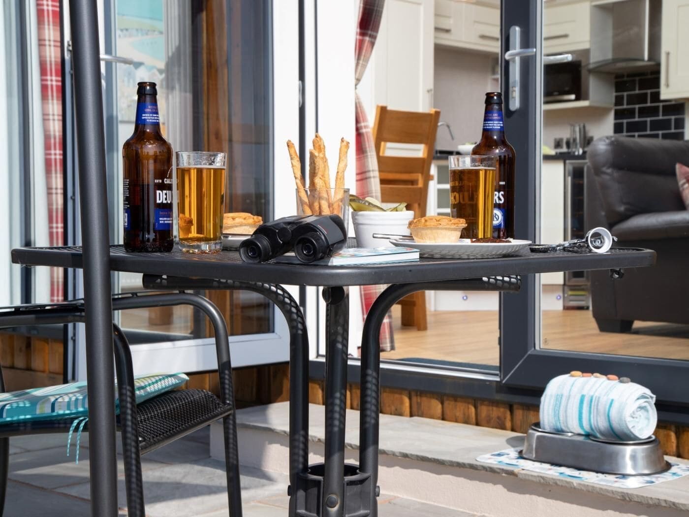 A table with beer bottles, glasses, baguettes, a camera, and a camera lens on a balcony with a view of the interior.
