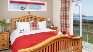 A bedroom with a wooden bed, a red blanket, a window with a red blind, a nightstand with a lamp, and a view of the sea. - 1 Bed in Castle Douglas (oc-rasc4) (Auchencairn)