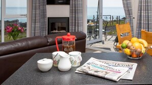 Un salón con un sofá de cuero, una mesa de centro con un periódico, un frutero y vistas al mar a través de amplios ventanales.