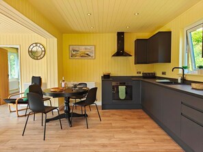 A modern kitchen with a built-in oven, a dining area with a round table and chairs, and a large window providing natural light. - 1 Bed in Conwy (oc-95098) (Ty'n-y-groes)
