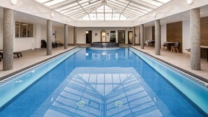 Ein Hallenbad mit Glasdach, Steinsäulen und einem zentralen Whirlpoolbereich.