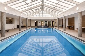 Ein Hallenbad mit Glasdach, Steinsäulen und einem zentralen Whirlpoolbereich.
