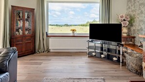 Ein Wohnzimmer mit großem Fenster, einem Holzschrank, einem Ledersofa, einem Fernseher auf einem Metallständer und einer dekorativen Vase.