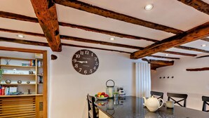 Una cocina con vigas de madera a la vista, un reloj, una encimera con una tetera y un comedor con sillas.