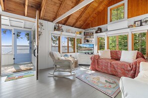 Ein Zimmer mit Holzbalkendecke, einem Schaukelstuhl, einer Couch und Blick ins Freie.