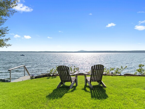 Two Adirondack chairs facing a body of water with a distant landmass, a wooden deck, and a grassy area.