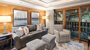 Una sala de estar con sofá, reposapiés, sillón, una mesa de centro, televisor y un ventanal con vistas al exterior.