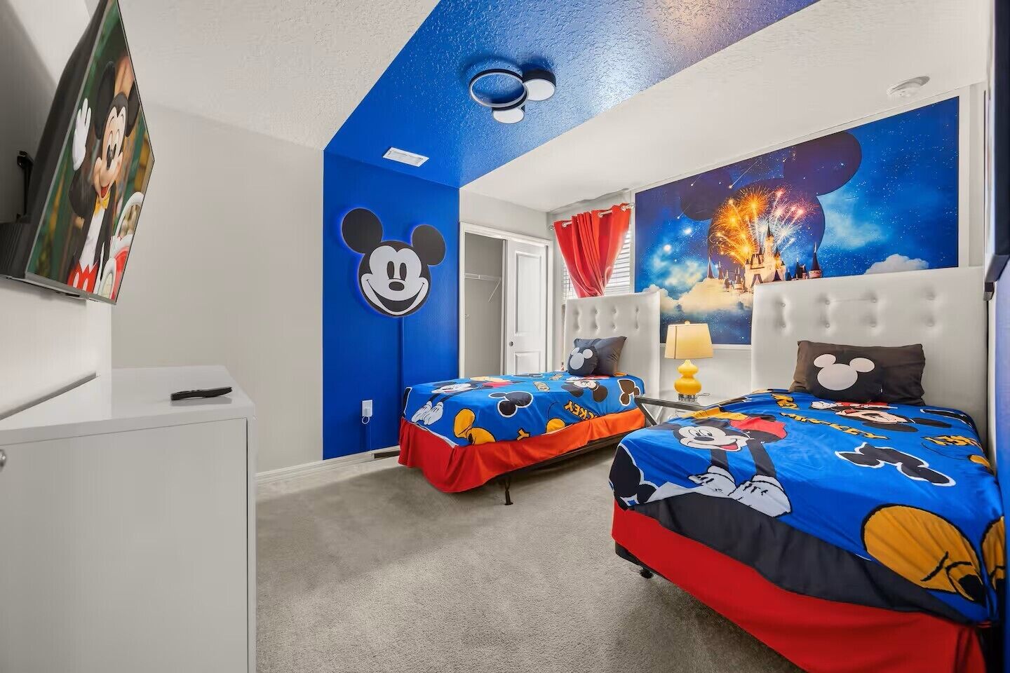 A room with two beds, a white dresser, a Mickey Mouse mural, and a large wall painting of fireworks.