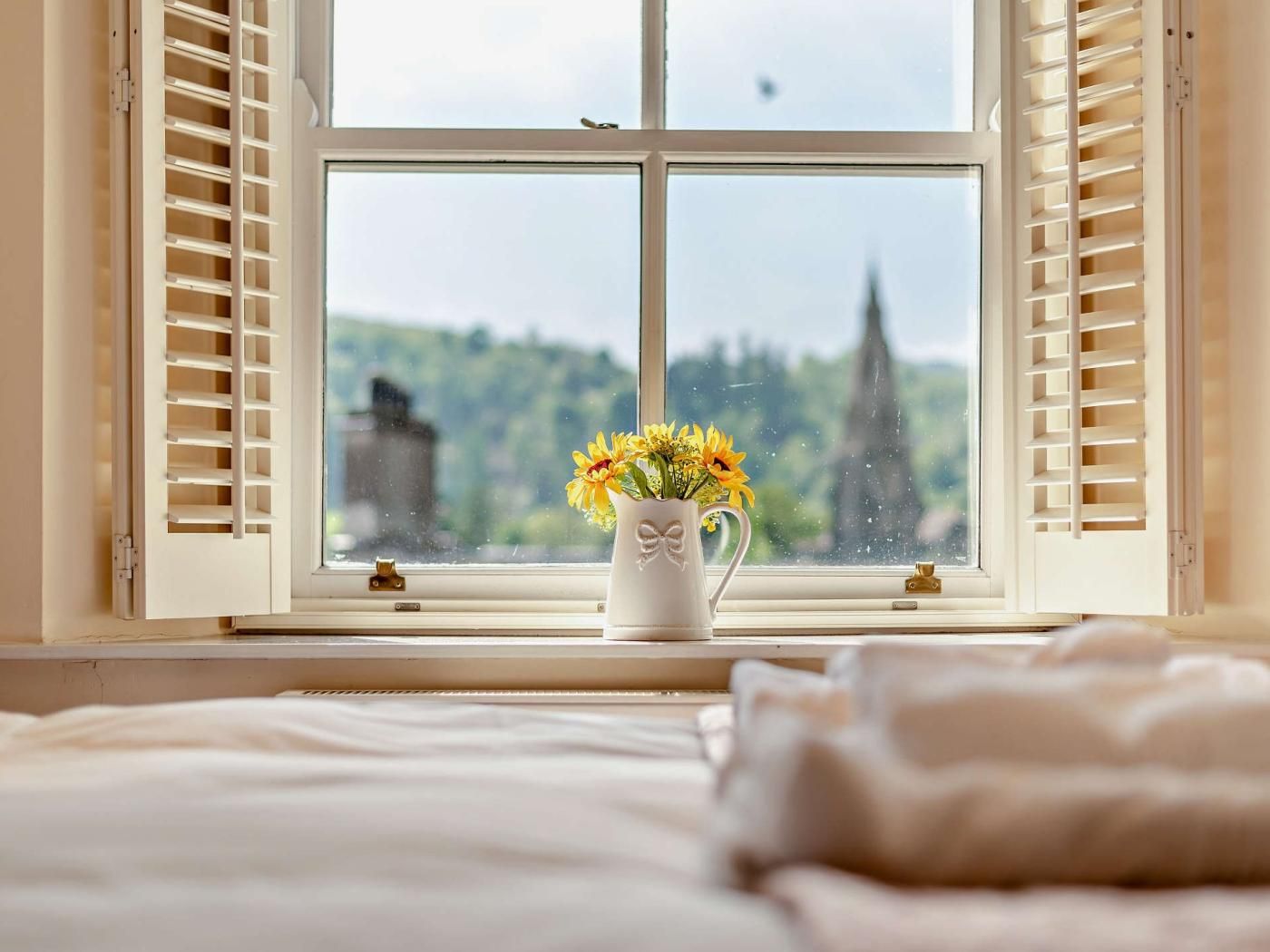 A window with open shutters, a vase with flowers, and a bed with a white cover.