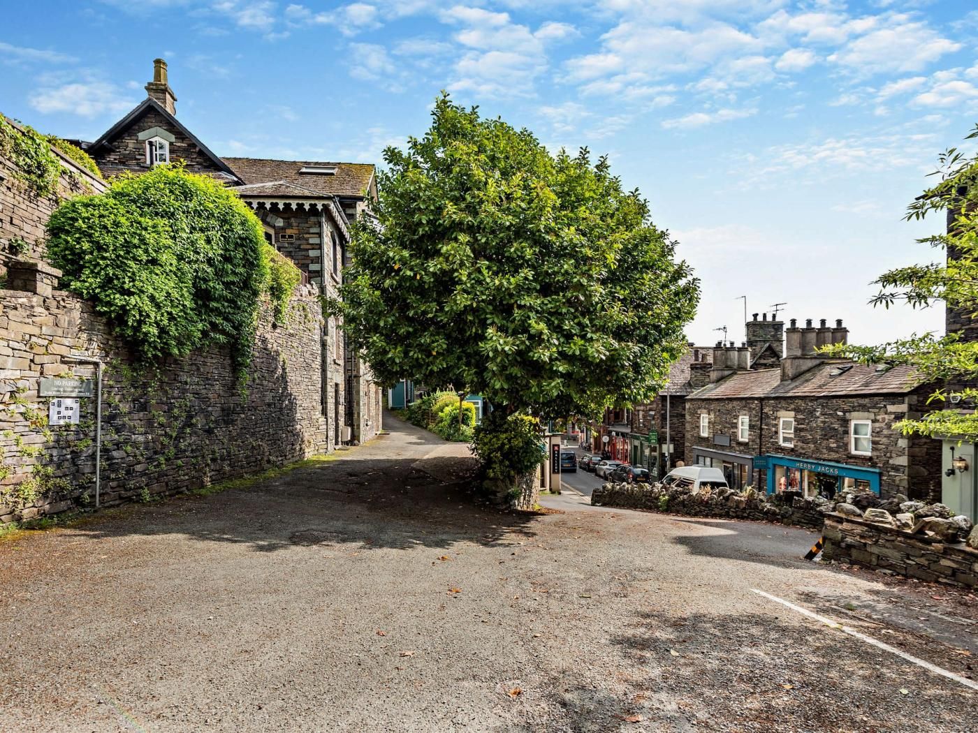 A narrow street with stone buildings, a tree-lined path, and a 'No Parking' sign.