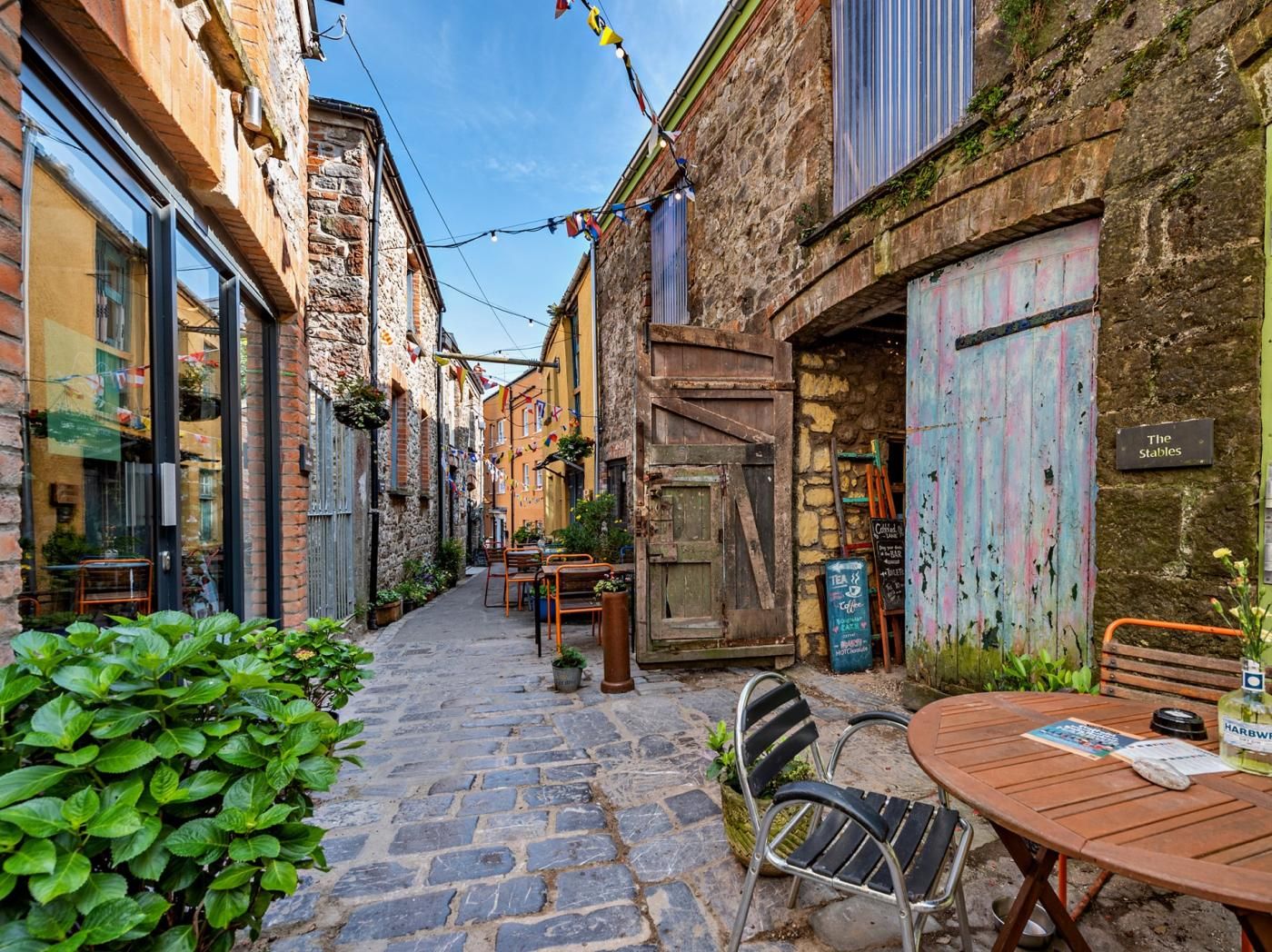 A narrow cobblestone street with buildings on either side, outdoor seating, and a sign for 'The Stables'.