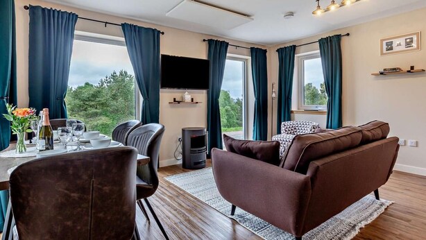 Ein modernes Wohnzimmer mit Sofa, Esstisch, Stühlen, Kamin und großen Fenstern mit Vorhängen.