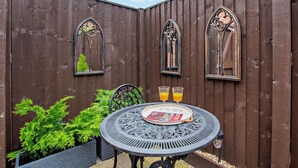 Eine Terrasse mit einem runden Metalltisch, zwei Stühlen und einem Holzzaun mit drei Rundbogenfenstern.
