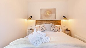 Ein ordentlich bezogenes Bett mit aufgerollten Handtüchern, zwei Nachttische mit Lampen und ein an der Wand hängendes Kunstwerk.
