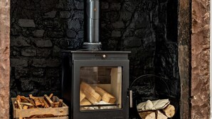 A wood-burning stove with a glass door, a stack of firewood, and a stone wall. - 2 Bed in Frizington (oc-95126) (Arlecdon)