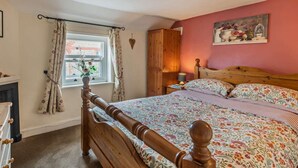 Une chambre avec un lit en bois, une literie à motifs floraux, une fenêtre avec des rideaux, une table de chevet avec une lampe et un tableau accroché au mur.