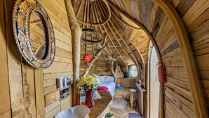 Un intérieur rustique en bois, avec un plafond voûté, un lit agrémenté d’un oreiller rouge, un banc en bois, un miroir et un vase rempli de fleurs.