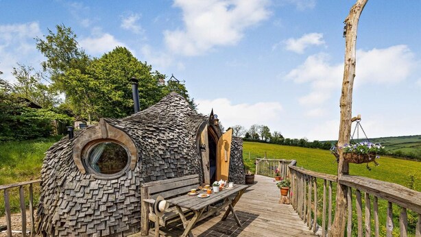 Une terrasse en bois avec une fenêtre circulaire, un banc et une table sur laquelle sont posés divers objets.