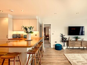 Eine moderne Küche mit einer Holzinsel, Barhocker, einem Fernseher und einer Wohnbereiches mit einer Couch und Pflanzen.