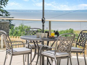 Une table ronde en métal, quatre chaises, un télescope et un verre de jus d’orange sont disposés sur un balcon donnant sur une étendue d’eau.