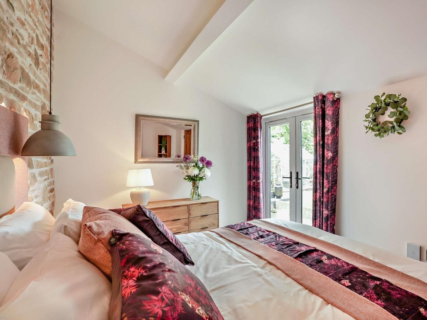 A bedroom with a bed, bedside table, lamp, mirror, window with curtains, and a wall-mounted wreath.