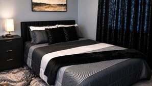 Un dormitorio con cama, mesita de noche, lámpara, un cuadro en la pared y cortinas estampadas.