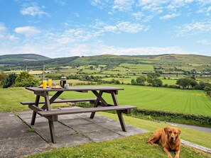 A picnic table with a dog, set against a rural landscape with rolling hills and houses in the distance. - 4 Bed in Isle Of Arran (oc-95184) (Blackwaterfoot)