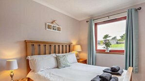 Un lit bien fait, avec des oreillers, une lampe de chevet, un petit tabouret en bois et une fenêtre avec des rideaux.