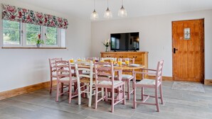 Un coin repas avec une table en bois, des chaises roses, un téléviseur et une fenêtre agrémentée d’un rideau à motifs floraux.