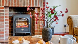 Una habitación acogedora con chimenea de ladrillo, una mesa con muffins y tazas, y un florero con flores rojas.