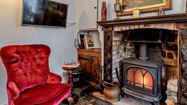 Una habitación acogedora con un sillón de terciopelo rojo, una chimenea con estufa de leña, un televisor montado y un cuadro enmarcados sobre la repisa.