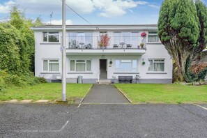 Ein zweistöckiges Wohnhaus mit Hauseingang, einem Balkon mit Sitzgelegenheit und einem großen Baum zur Rechten.
