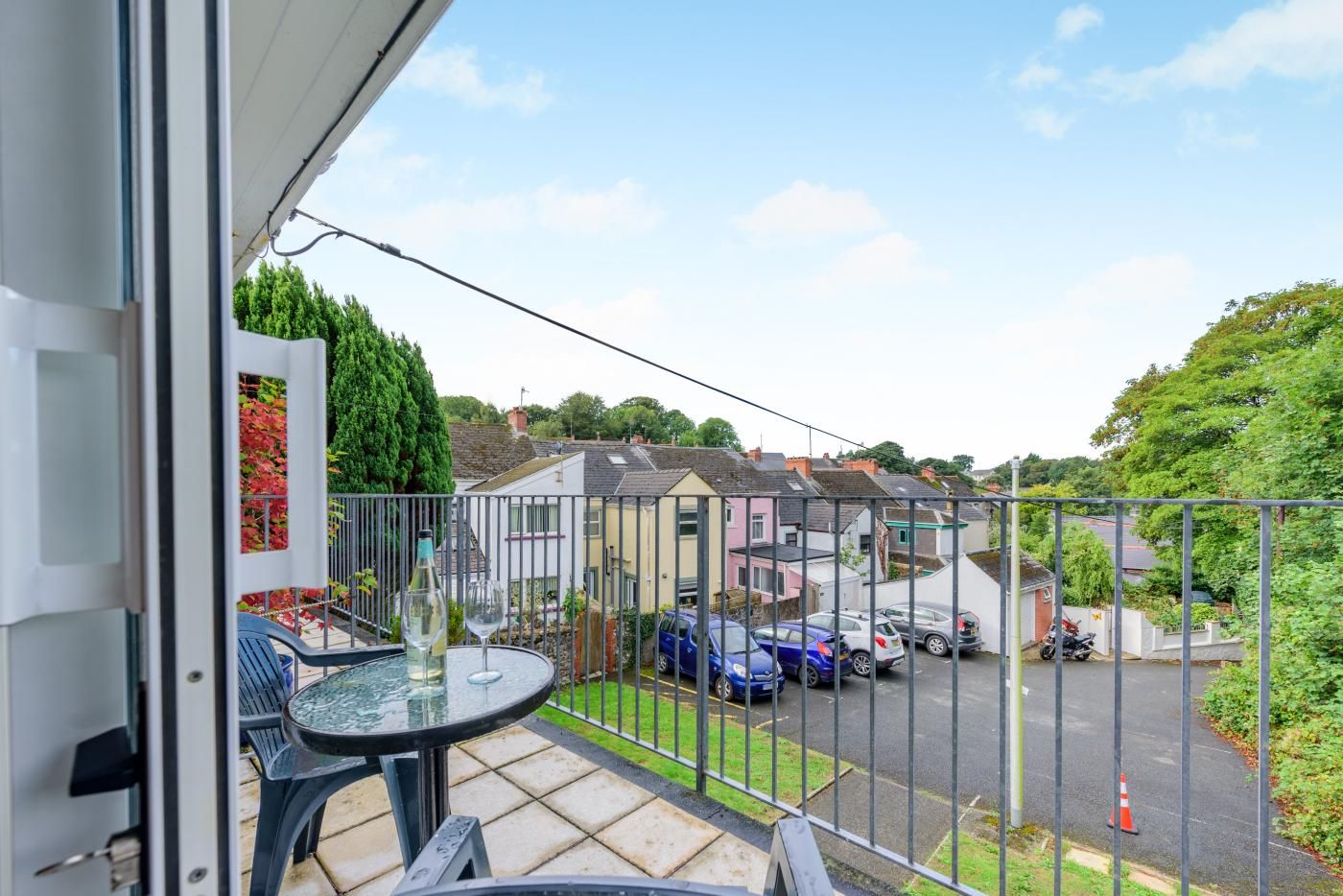 A balcony with a table and chairs, overlooking a parking area and houses.