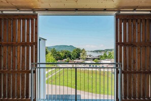 A view from a balcony with a metal railing, overlooking a grassy area, a body of water, and distant buildings. - Apartement with lake view (Unterburg am Klopeiner See)