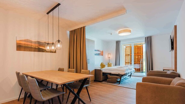Moderni hotellihuone, jossa on puinen ruokapöytä, mukavat istuimet, sohva, sänky ja suuri ikkuna, jossa on verhot.