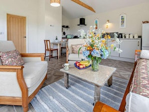 A living room with a sofa, a coffee table with fruit and flowers, a dining area with a chair, and a kitchen in the background. - 1 Bed in Liskeard (oc-95182) (Looe)
