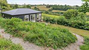 Ein kleines Gebäude mit dunklem Dach und weißen Fensterrahmen, umgeben von üppiger Vegetation und einem Feldweg.