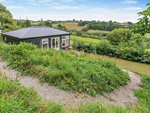A small building with a dark roof and white-framed windows, surrounded by lush greenery and a dirt path. - 1 Bed in Liskeard (oc-95182) (Looe)